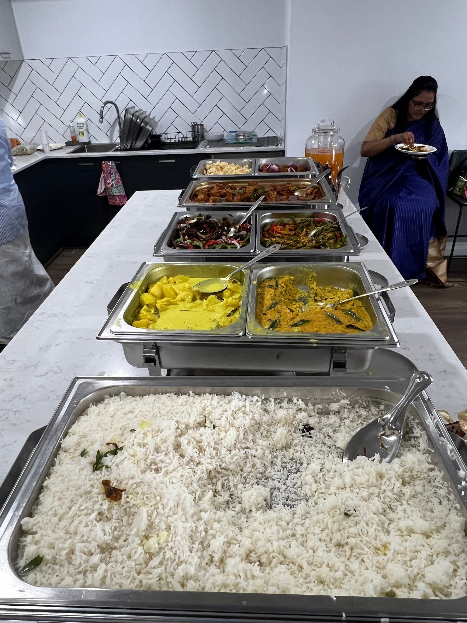 Woman in blue sari serving food at a Sri Lankan catering buffet setup with chafing dishes