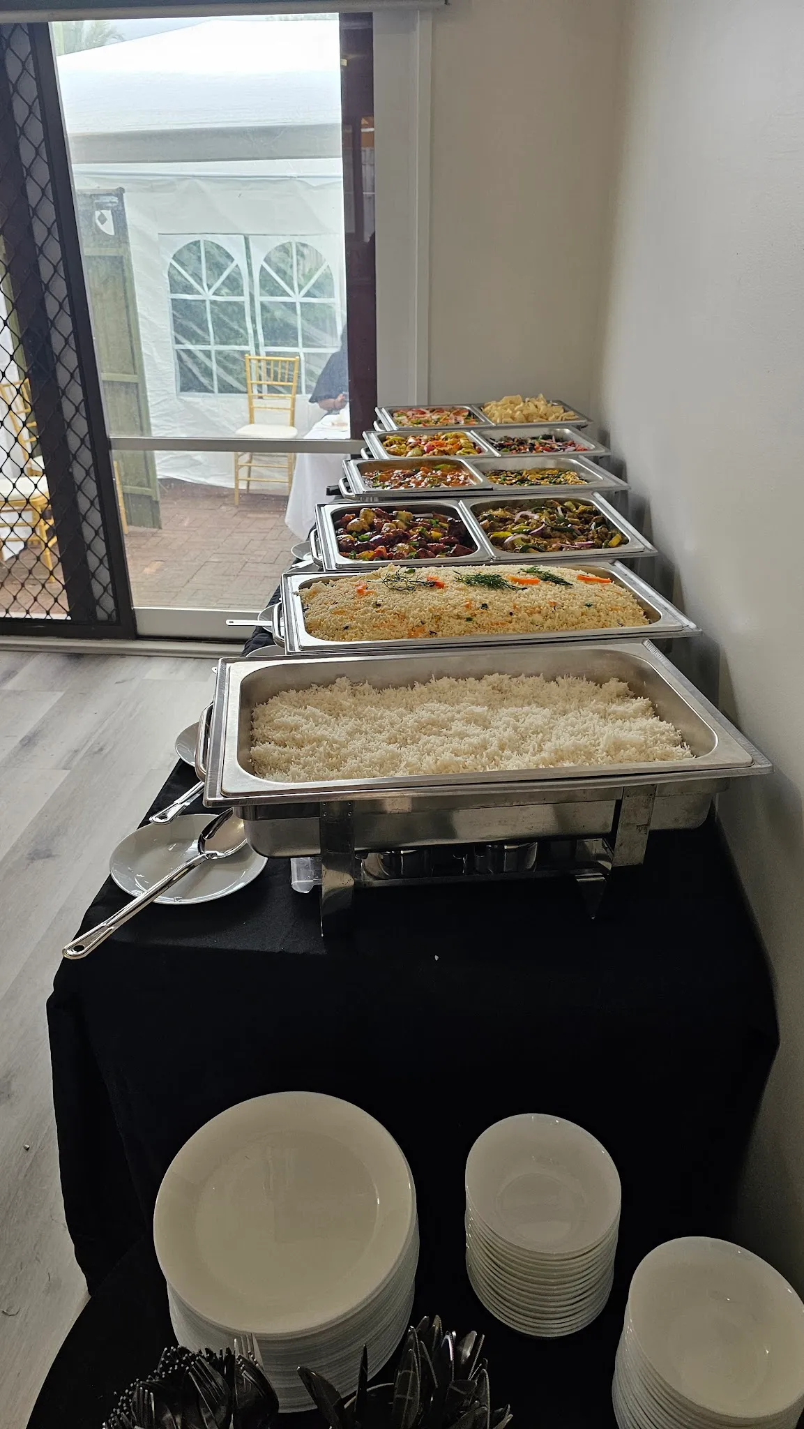 Sri Lankan buffet station with stacked plates and chafing dishes, window view of outdoor marquee tent