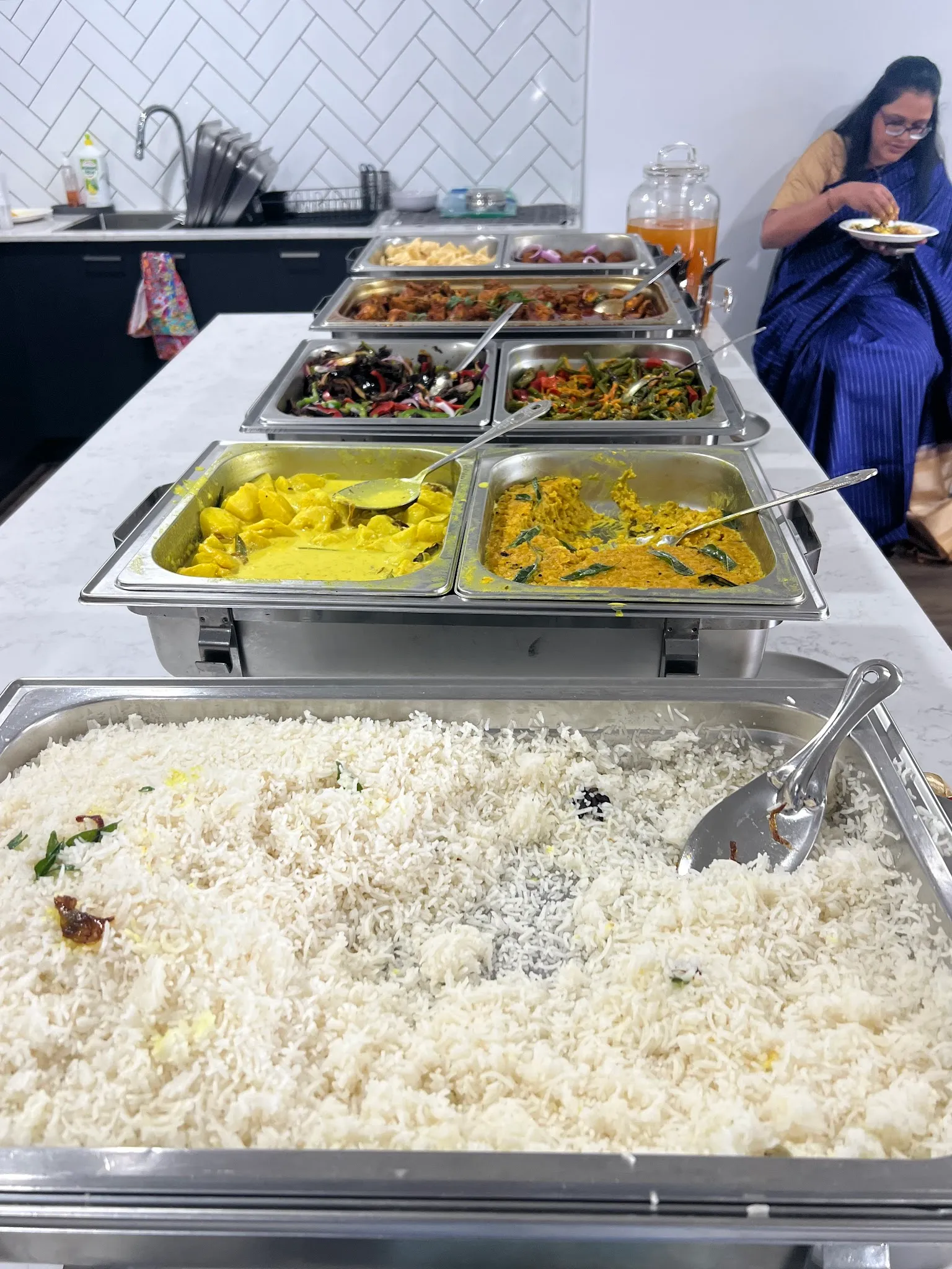 Home kitchen catering setup with chafing trays and woman in blue sari serving food